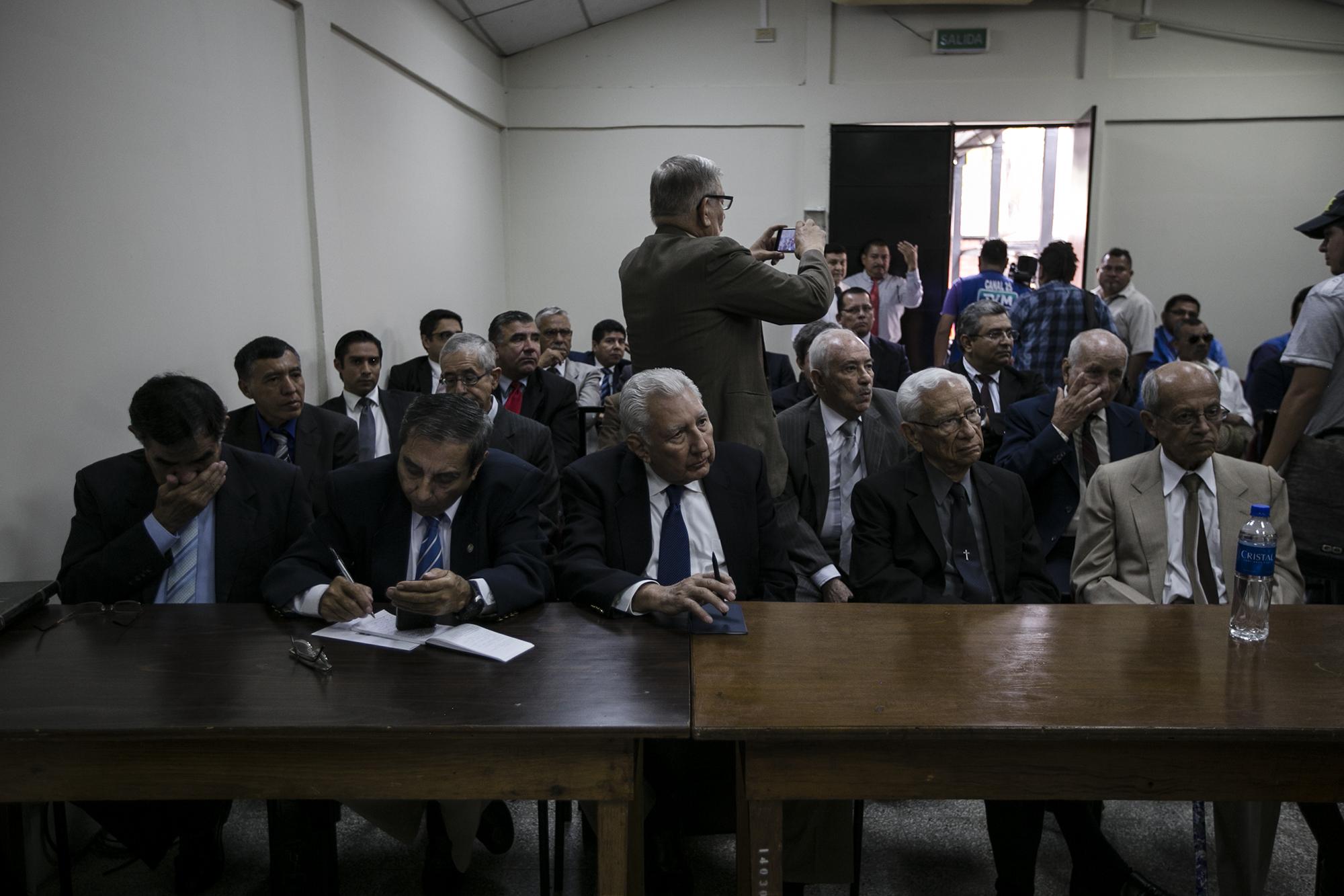 Militares acusados por la masacre de El Mozote, minutos antes de iniciar la audiencia de intimación en Gotera. En primera fila, de izquierda a derecha: Juan Antonio Rodríguez Molina, José Mario Godínez Castillo, Rafael Flores Lima, José Guillermo García y Luis Adalberto Landaverde. Atrás de ellos se encuentra, de pie, fotografiando a los periodistas, el coronel retirado Natividad Jesús Cáceres, quien fue jefe operativo del BIRI Atlacatl y tercero al mando en la masacre. Foto: Fred Ramos