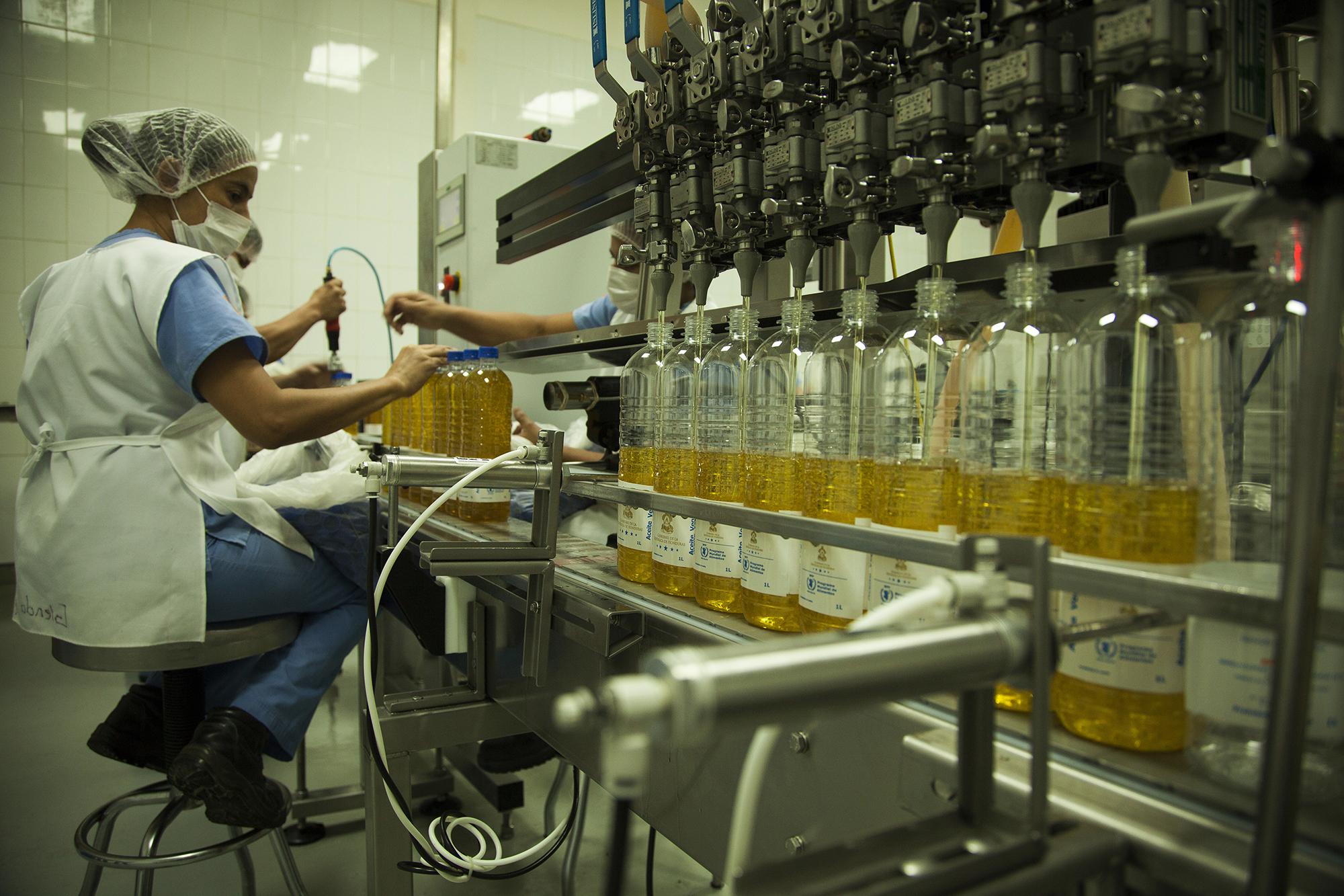 En la planta de envasado de Hondupalma, los trabajadores llenan botellas de aceite con el logo de la República de Honduras. Son los productos para regalías que utiliza el gobierno, que se ha convertido en uno de los principales impulsores de la producción derivada de la industria de la palma africana desde 1971, cuando se determinó la siembra de palma africana como una política de estado en todo el Valle del Aguán.
