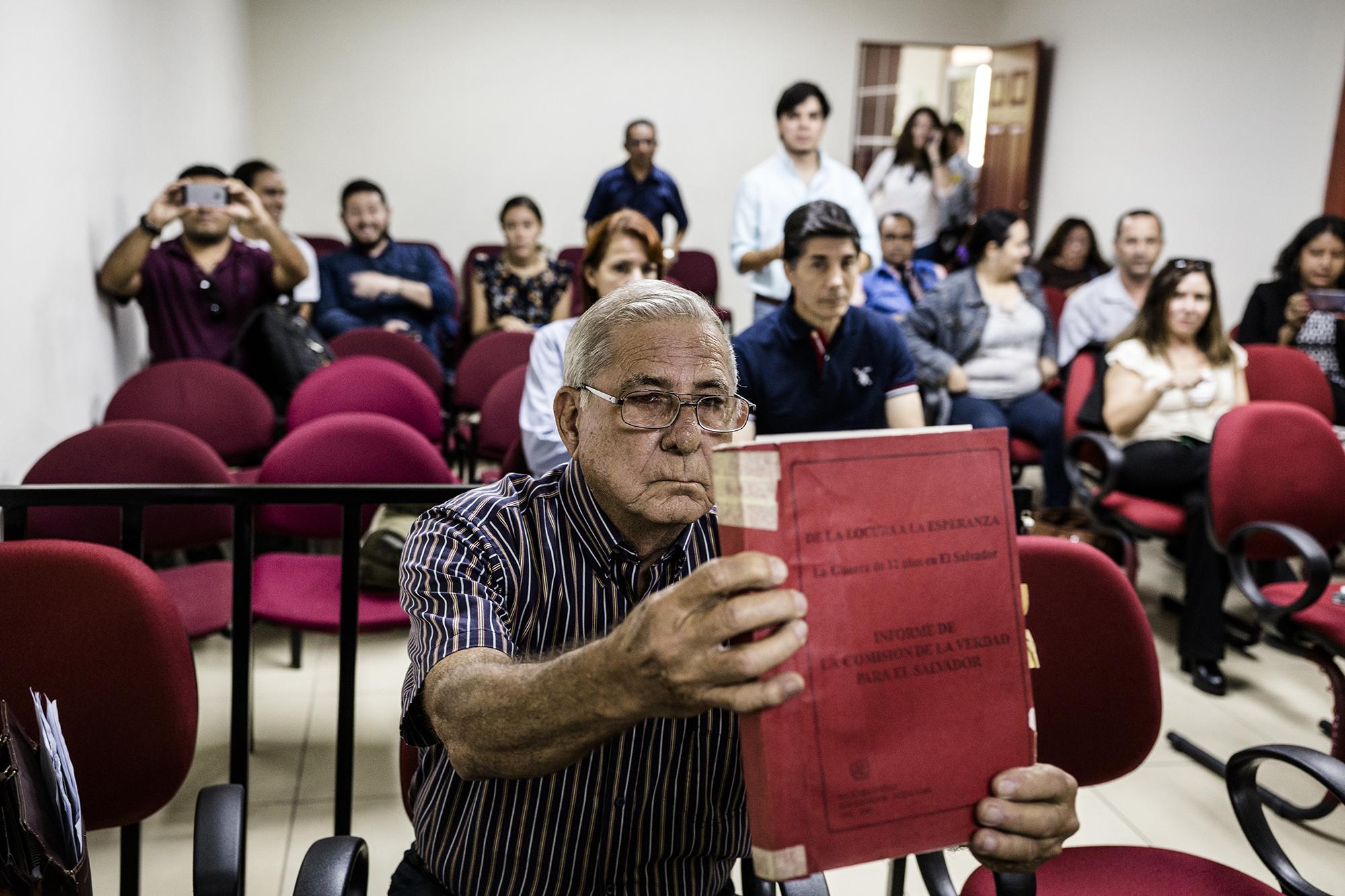 El general Juan Rafael Bustillo, sentado en el banquillo de los acusados, muestra un ejemplar del informe de la Comisión de la Verdad , en el que se narran los casos ejemplificantes de violaciones a los derechos humanos cometidas en la guerra civil salvadoreña (1980-1992). Bustillo llegó el 19 de octubre de 2017 al Juzgado de Instrucción de San Francisco Gotera para responder, al igual que otros 17 militares, por la masacre de El Mozote y caseríos aledaños, en la que participaron diversas unidades del Ejército salvadoreño y en el que se asesinó a alrededor mil campesinos en la zona norte del departamento de Morazán, en diciembre de 1981. Foto: Fred Ramos