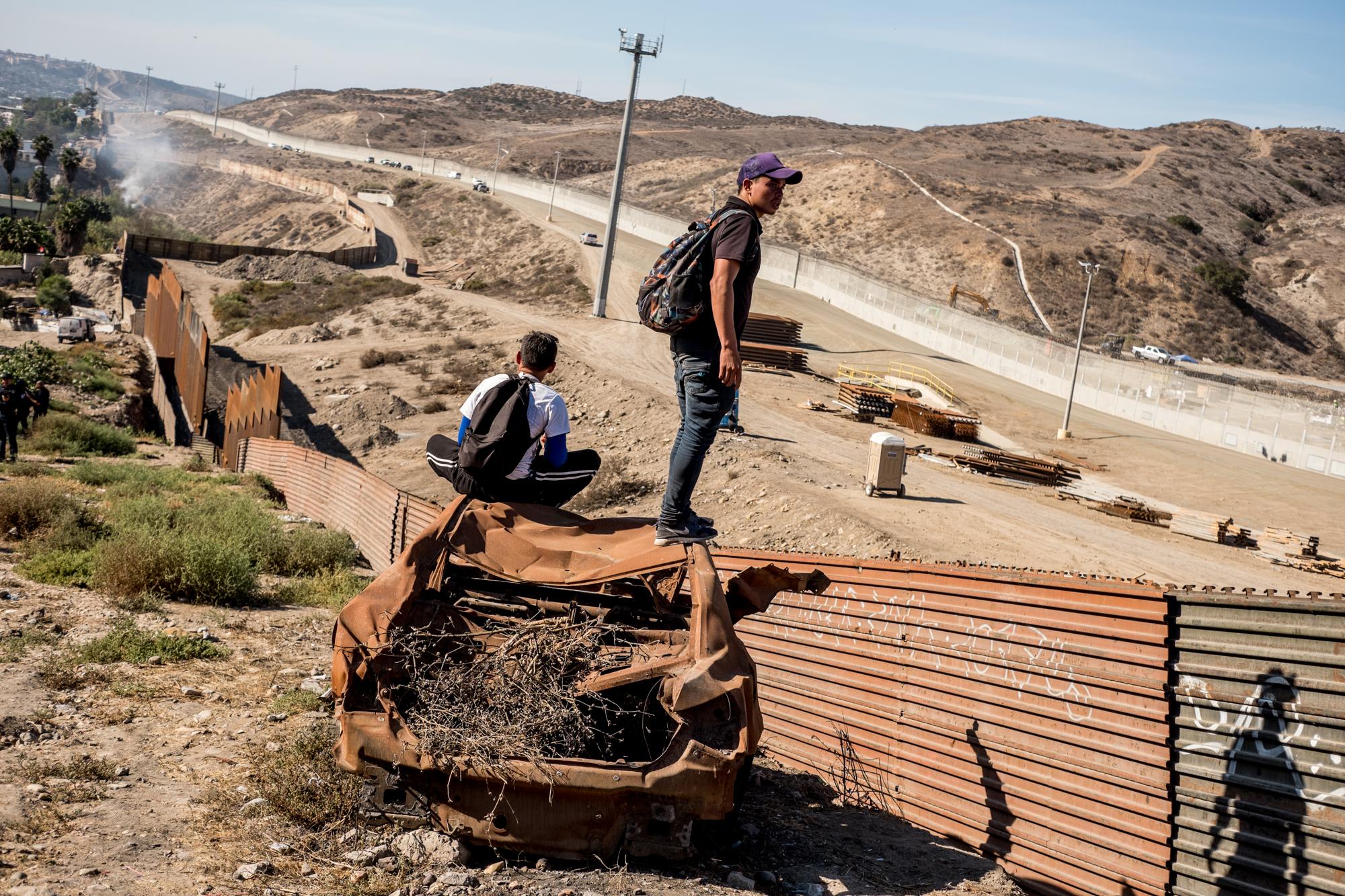 Migrantes hondureños observan hacia el territorio estadounidense. Ese espacio entre la barda oxidada y el muro blanco del fondo es utilizada por la Patrulla Fronteriza para recorrer en vehículo la frontera, pero ya se considera territorio estadounidense. Al fondo de la imagen, una nube de gas pimienta lanzada por helicópteros de la Patrulla Fronteriza de Estados Unidos repliega a los migrantes que venían atrás, en un grupo mucho más numeroso.   