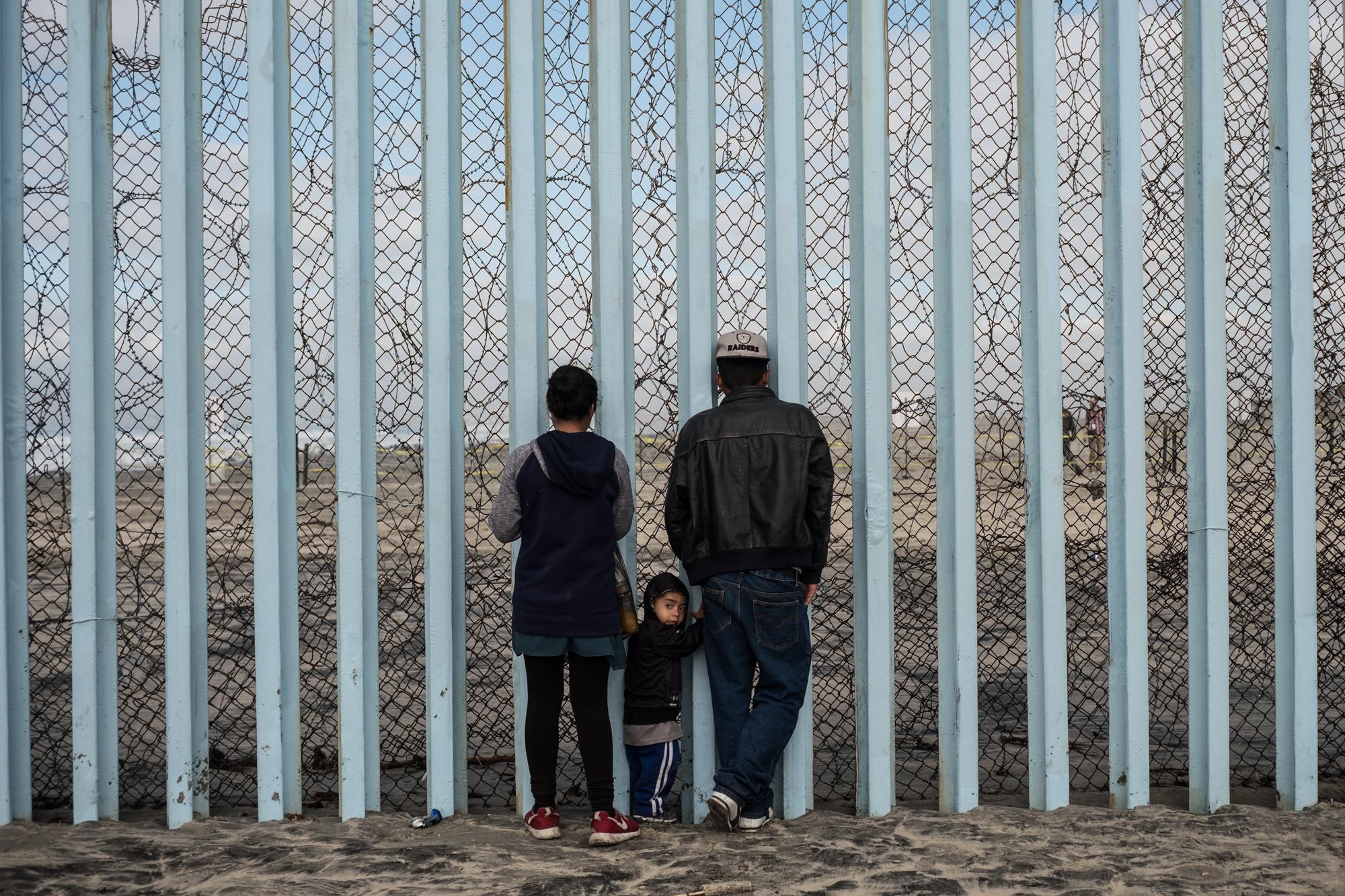 Nicole Cruz y David García observan hacia el lado estadounidense del muro que divide México y Estados Unidos, junto a su hijo de dos años, Mateo García. La familia García decidió huir de Honduras por las amenazas recibidas de la pandilla Barrio 18. Foto archivo El Faro.