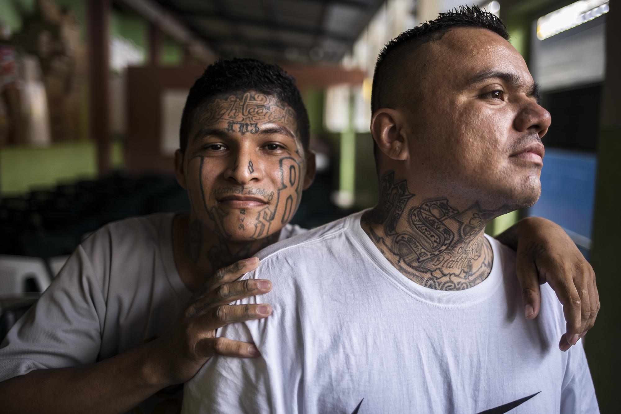 Sonríen, se abrazan, algo impensable para ellos mismos unos años atrás. Irvin Rivas, de 28 años (izquierda), y Dani José Parada, de 32, salieron del estudio bíblico para conversar con El Faro, en un salón de clases del sector seis. Irvin se brincó al Barrio 18 Revolucionarios, de la colonia Valle del Sol, en el municipio de Apopa, y con los números de su pandilla tatuó su cara. Cumple una pena de de diez años por el delito de extorsión. Dani está procesado por asociaciones ilícitas cuando era miembro de la MS-13, del sector de Mariona, la colonia que rodea el penal La Esperanza. Tatuó en su cuello las letras de su organización. 