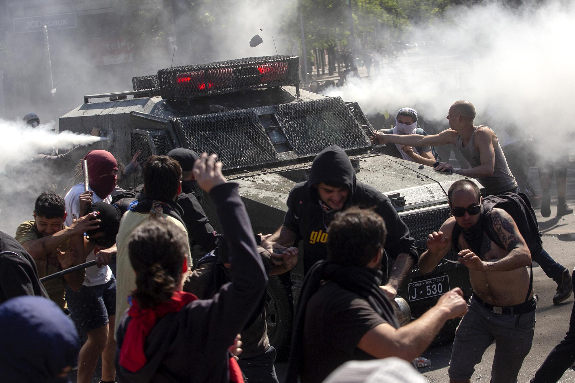 Estallido social en Chile. El viernes 18 de octubre, el aumento en casi 4 centavos de dólar en la tarifa del metro provocaron una oleada de protestas en Chile que han provocado 18 muertos, cinco de ellos por acción de agentes del estado, en medio de crecientes denuncias de abuso policial y militar. Las maifestaciones se han convertido ahora en una suerte de catarsis nacional sin final a la vista y los chilenos también protestan por las bajas pensiones, precariedad en salud y educación, una desigualdad crónica o la poca empatía de la clase política. En la imagen, manifestantes se enfrentan con un vehículo de la policía antidisturbios el 20 de octubre. Foto por Claudio Reyes /AFP.