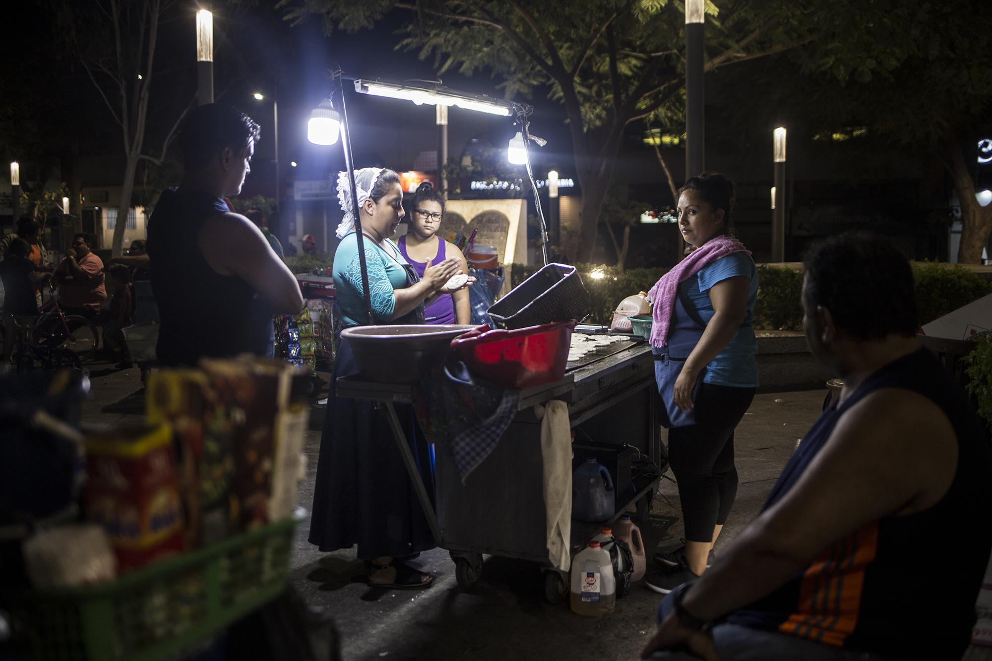 En medio de la Plaza Barrios, en el Centro capitalino, hay una pupusería rodante. Es una armazón de lámina, que se ilumina con una batería de corriente directa. Son casi las diez de la noche, y Blanca sigue en el negocio. Las ventas no fueron como la de todos los sábados. La afluencia de gente ha sido menor en el Centro Histórico de San Salvador, y algunos vendedores hacen esfuerzos como este. Para Blanca no es opción dejar de trabajar. 