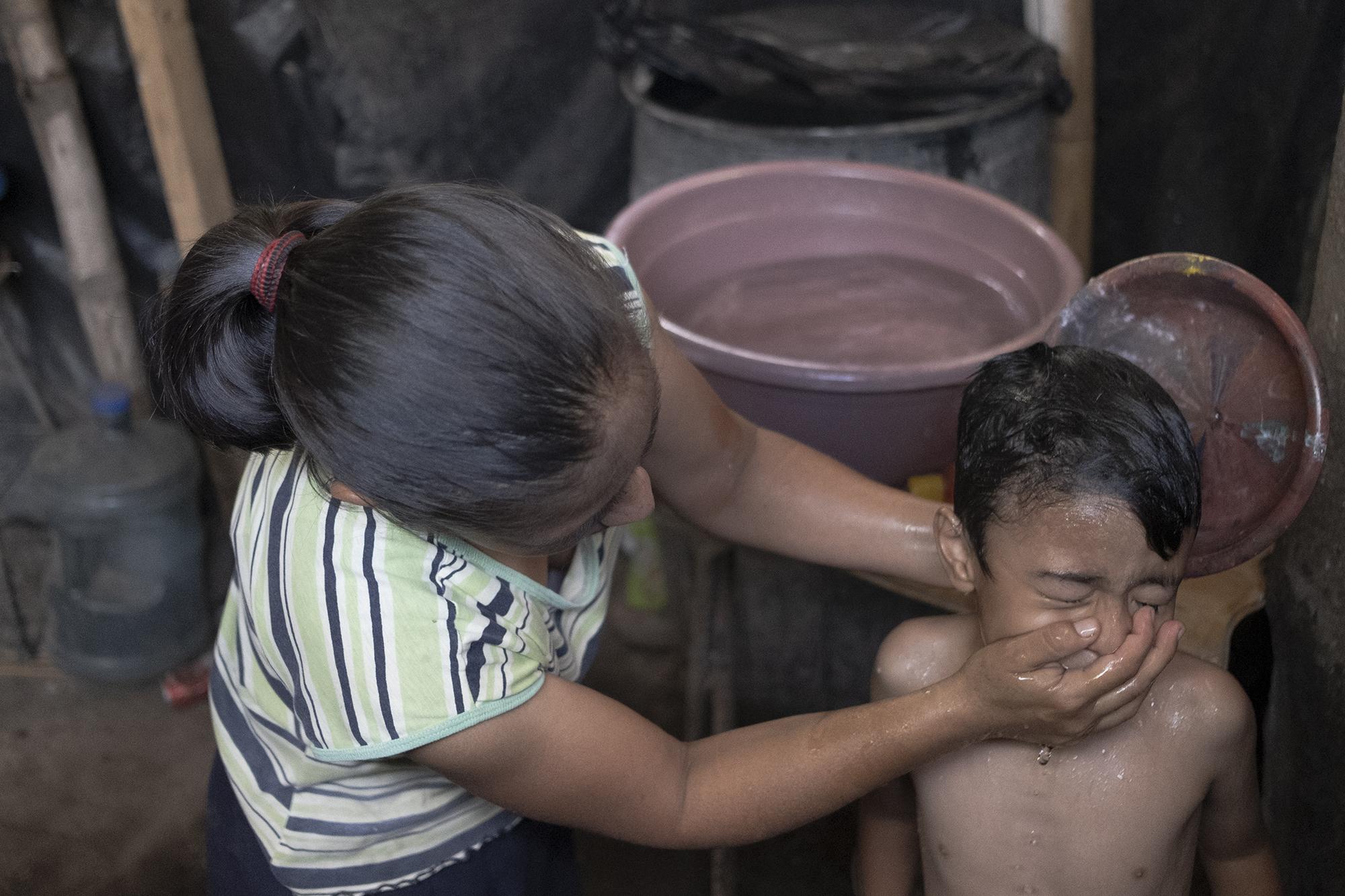 Raquel Flores, de 41 años, da un baño a su hijo Gabriel, de 5. Aquí no hay agua en las casas, deben acarrearla todos los días desde chorros comunitarios. Raquel no cuenta con un trabajo formal. Antes de la cuarentena hacia trabajo de costurera para los vigilantes del Sistema de Integración Centroamericano, pero en su mayoría ya no llegan a laborar. En la casa de Raquel nadie salió beneficiado con el bono de $300. 