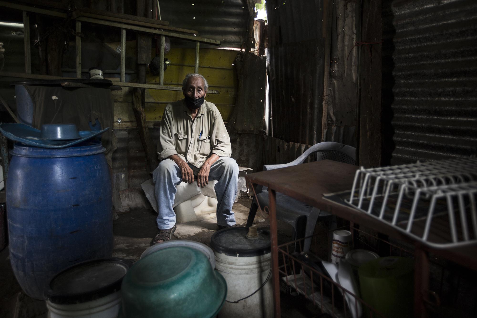 Mario Quintanilla trabajaba como mecánico automotriz. Ahora es un hombre solo, sin sus hijos, sin su esposa y sin trabajo. Desde que empezó la cuarentena, la caridad de sus vecinos le permite sobrevivir. Tiene 80 años y vive entre un cúmulo de repuestos viejos y bajo láminas agujereadas por donde las aguas lluvias se cuelan. Foto de El Faro: Víctor Peña.