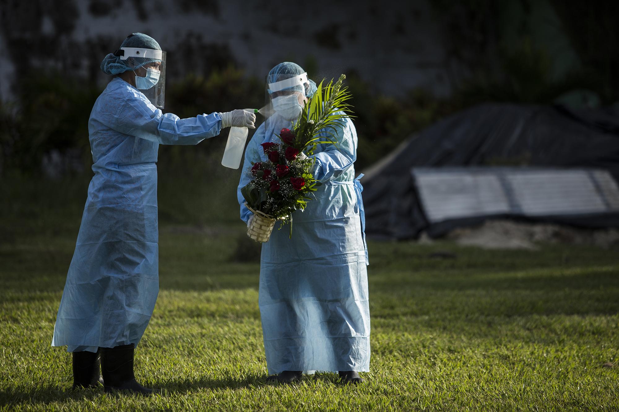 Empleados del cementerio desinfectan la ofrenda floral que llevó una pareja que asistió al entierro de una persona fallecida en el hospital Militar de San Salvador. Los dolientes no pueden ni siquiera colocar las flores en la tumba, sino que esa labor la hacen los empleados. Foto de El Faro: Víctor Peña. 