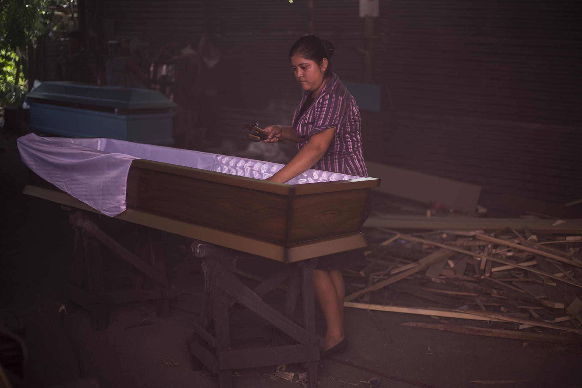 Xiomara Cruz es la única mujer en el taller, y también la única que trabaja en el área de tapicería y acabados. Xiomara aprendió este oficio con sus padres, pero asegura que es más rentable trabajar en un taller ajeno a su familia. A diario tapiza diez ataúdes, al menos en los últimos meses, un trabajo que le deja ganancias de $40 por día. Ella y su hermano pagan seis dólares diarios para llegar a su trabajo desde el cantón Buenos Aires, del municipio de San Pedro Perulapán. Por esta caja en la que trabajará una hora, Xiomara gana $5.