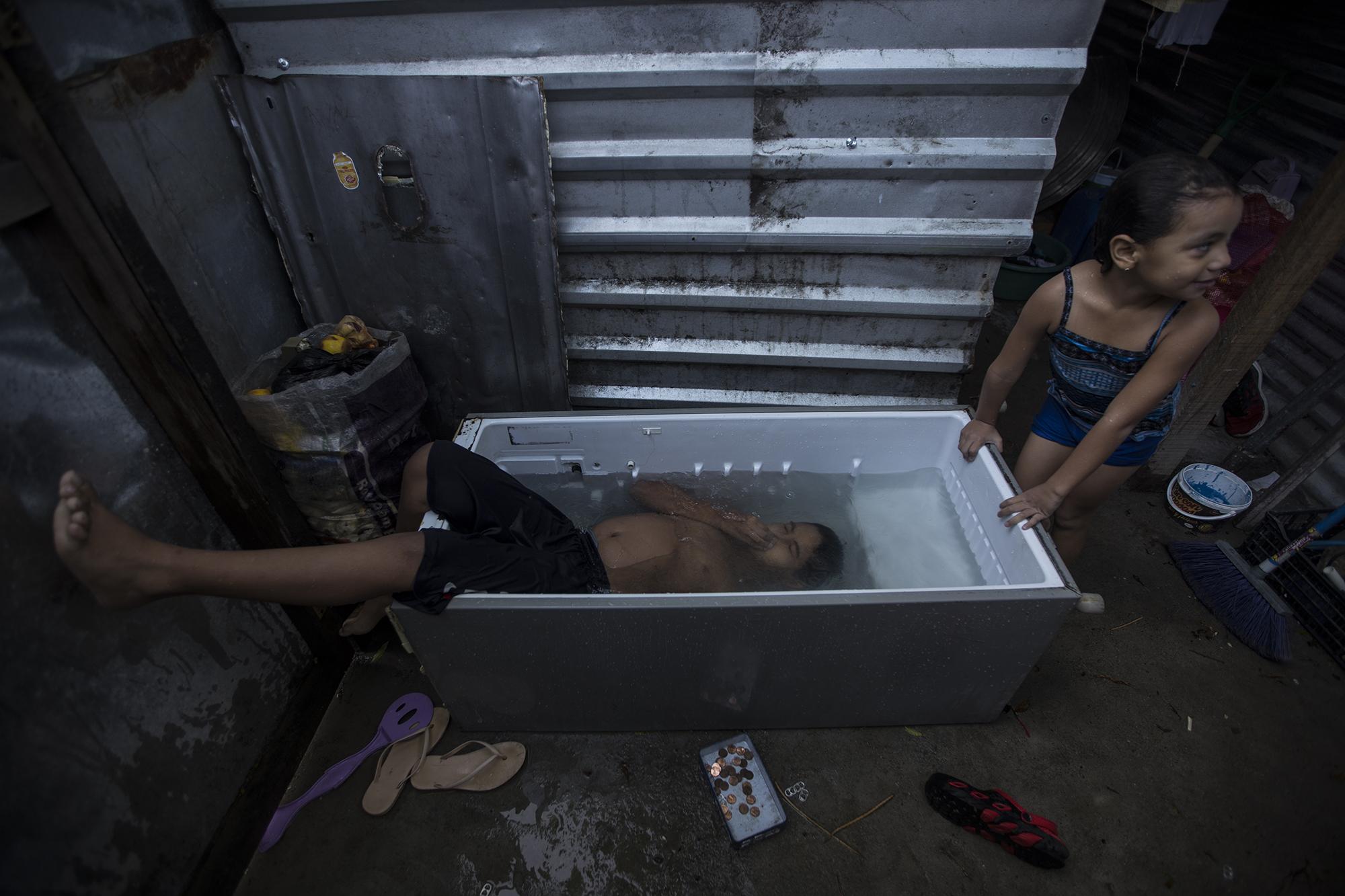 16 de mayo de 2020. Pedro Montano, de 11 años, se sumerge en los escombros de una refrigeradora transformada en pila. Milagro, su hermana, espera su turno al interior de su vivienda en la comunidad 15 de Marzo. La casa fue construida con desechos que sus padres recogieron en la calle. Cada día, estos niños tienen derecho a una hora de recreación en su 