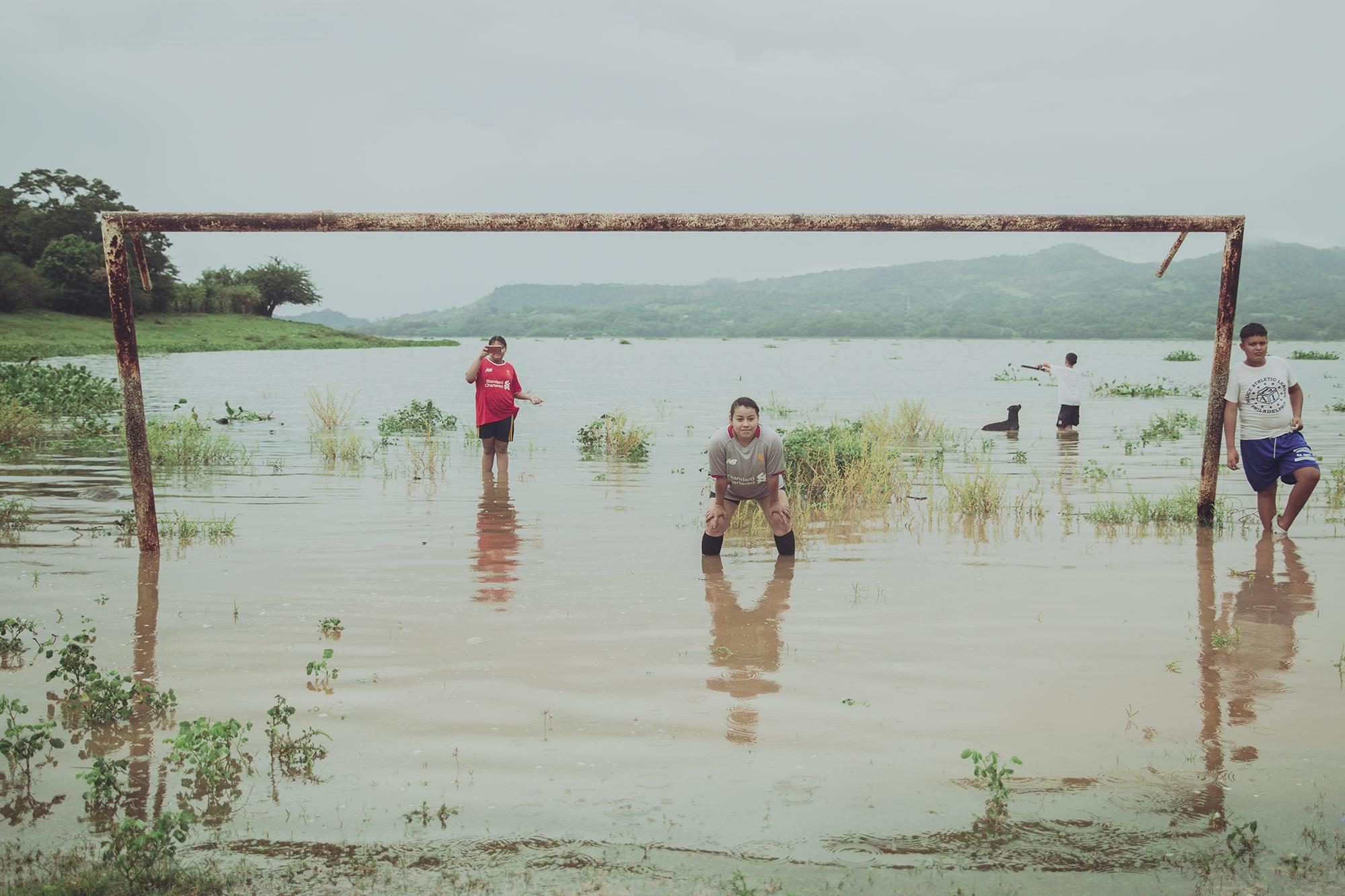 En Suchitoto había llovido toda la tarde del jueves 27 de agosto. La tormenta, según los meteorólogos, era influencia directa del huracán Laura que había golpeado las costas de Estados Unidos. A los lugareños poco les importaba la lluvia, menos a los que participaban en un partido de fútbol a las orillas del lago Suchitlán, en Suchitoto, a poco más de 47 kilómetros de San Salvador. Si el coronavirus no detuvo los partidos cantonales, menos lo hará una tormenta. El partido que se jugaba era importante, reunía a dos equipos de cantones con rivalidad futbolística, separados por 5.6 kilómetros. La portería del lado oeste era defendida por el equipo del cantón Aguacayo, que vestía el uniforme azulgrana del Barcelona. Era el equipo visitante. Las locales, del cantón San Juan, vestían una réplica del uniforme rojo del Liverpool. En la cancha, por no decir potrero, se lucía toda la ruralidad del encuentro. Algunas jugadoras iban descalzas y marcaban sin ningún temor a las que utilizaban zapatillas para fútbol. La inusual cancha tenía un desnivel hacia el noreste. El “engramillado” es una mezcla de zacate, lodo y excremento de vaca. Elda Recinos chapoteaba en el agua cuando aún caían algunas gotas. Para caminar, bajo la portería, la arquera de 18 años tenía que hacer un esfuerzo mayor que el de sus compañeras. El agua le llegaba hasta los tobillos y, en algunos puntos de su arco defendido, incluso hasta las rodillas. Cuando Elda se tiraba para cazar la pelota, más bien parecía que se aventaba un clavado. 