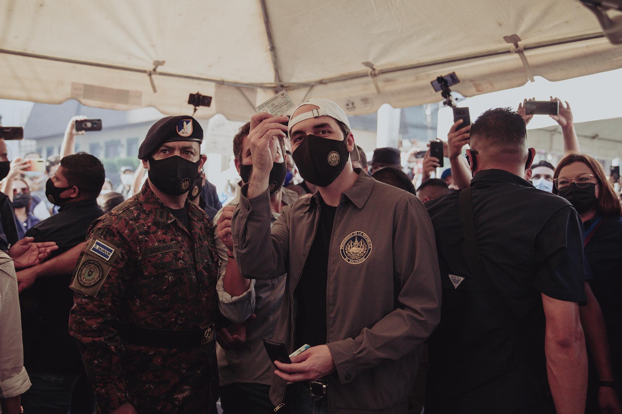 Nayib Bukele muestra su DUI antes de caminar hacia la JRV 502 donde ejercería su voto. El presidente llegó al centro de votación Bulevar del Hipódromo entre música y rodeado de la seguridad presidencial y decenas de personas particulares y de medios de comunicación. Foto de El Faro: Carlos Barrera