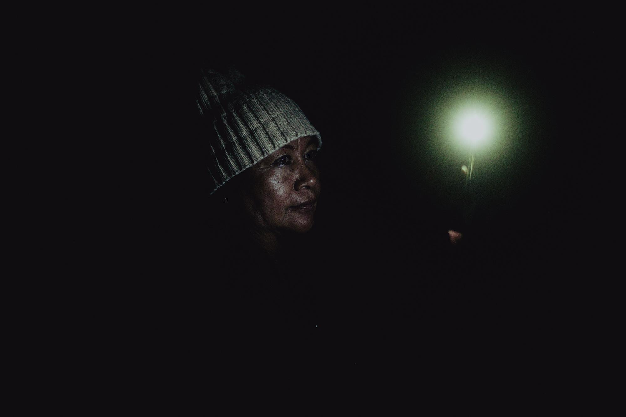 Victoria Panameño, de 58 años, trata de iluminar con la lámpara de su celular un sector del inmueble de la ex fábrica Florenzi durante su turno de vigilancia la noche del 15 de marzo de 2021. Según comentó, esa noche el lugar lo cuidaron para que nadie se robara nada, ya que de eso depende la indemnización que les corresponde por los años laborados. La extrabajadora le dedicó treinta años de su vida a la fábrica y con el pago que reciba piensa montar un negocio porque debido a su edad se le complica conseguir un trabajo en otra fábrica. Foto de El Faro: Carlos Barrera.