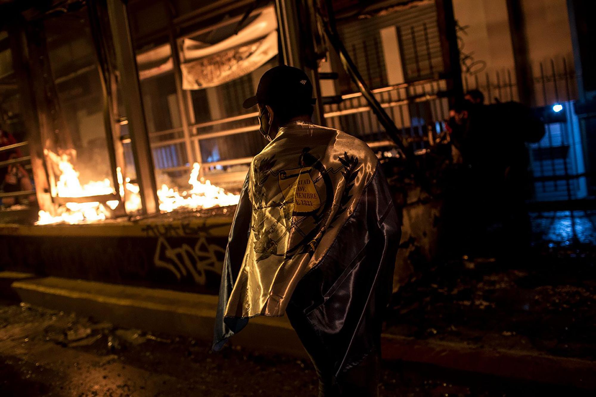 Un manifestante, con la bandera nacional en las espaldas, pasea frente a la parada del Transmetro de la novena calle, entre 6 y 7 avenida, en llamas durante los disturbios entre policías y manifestantes por la aprobación del presupuesto 2021. Foto: Simone Dalmasso/Plaza Pública