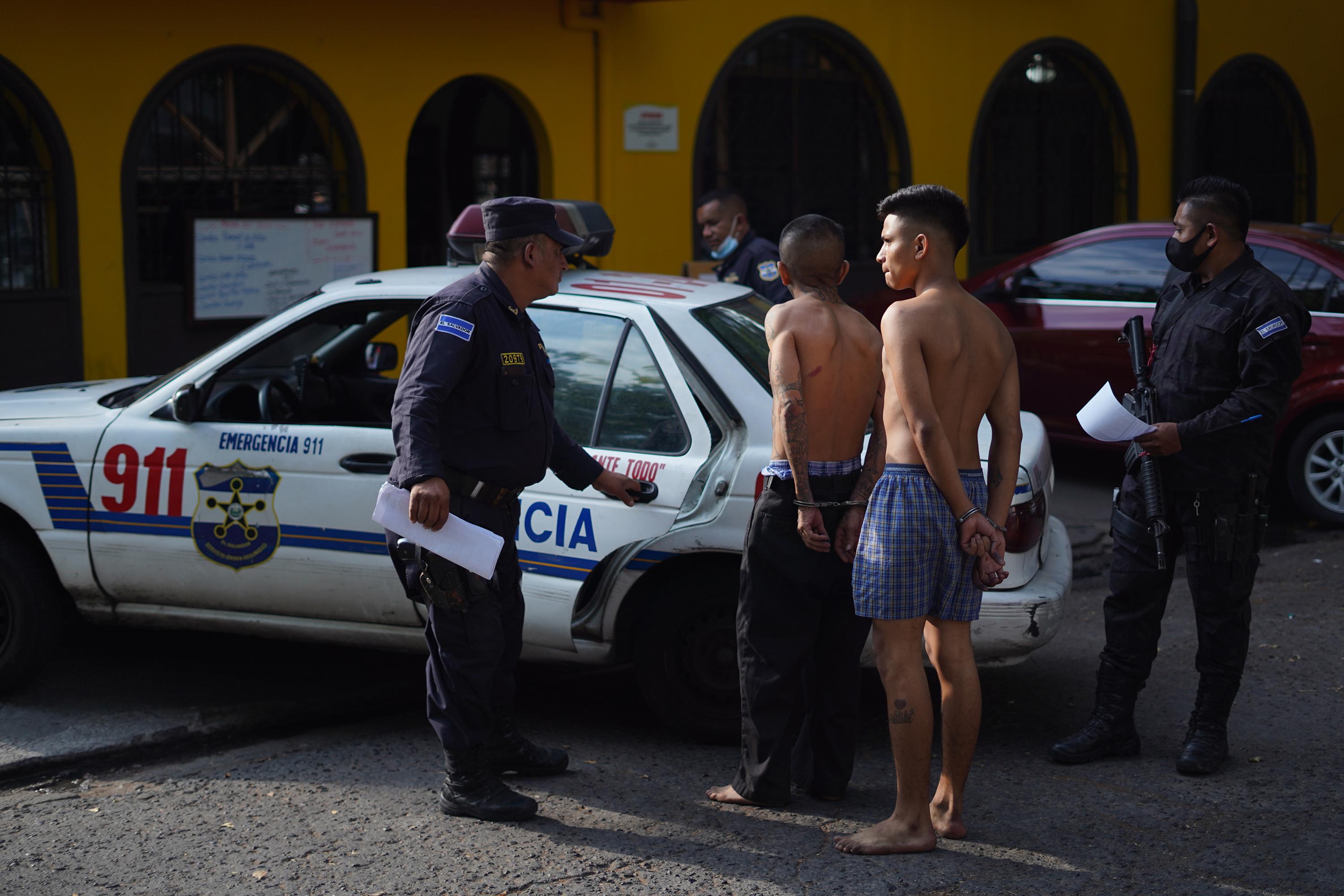 Agentes de policía suben a dos detenidos a una patrulla frente al edificio de la Procuraduría General de la República, en San Salvador. Los supuestos pandilleros fueron capturados bajo el régimen de excepción y llegaron a la Procuraduría para buscar representación legal de un defensor público. Foto de El Faro: Víctor Peña.