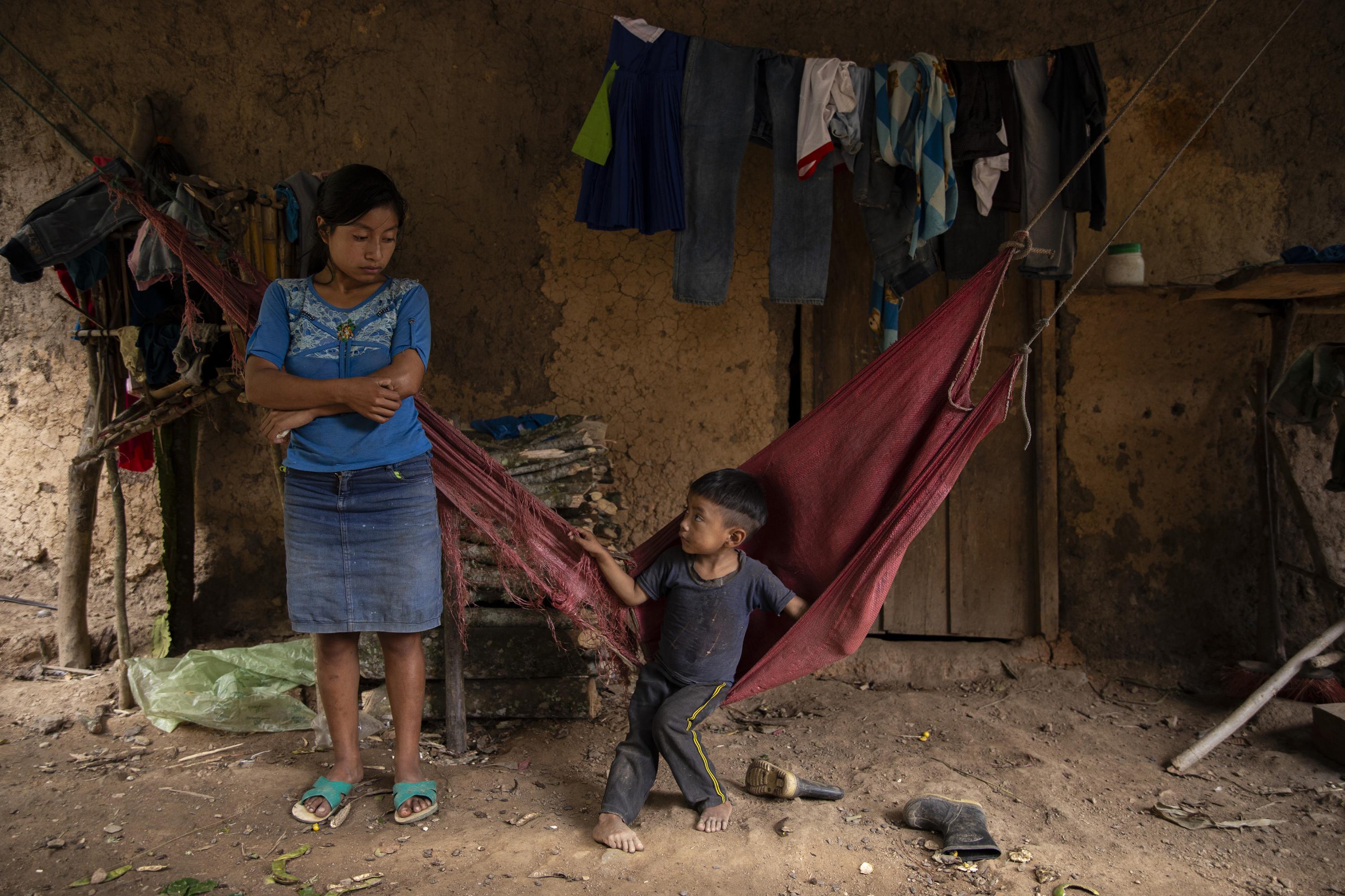 Herminia Gutiérrez y su hijo viven en la misma casa donde murieron Adalicia, de cinco años, y María Isabel, de seis meses, en septiembre de 2021. Sus hermanitas son parte de la tasa de mortalidad de 18 menores que la comunidad ha registrado desde el año 2013 a causa de la desnutrición. La vivienda está construida en medio de una ladera, sin acceso a agua potable, rodeada de cafetales, patos, gallinas y algunos perros desnutridos que duermen sobre el patio de tierra. Herminia ahora tiene 20 años, y sólo alcanzó una altura de 1.25 metros. Creció bajo pobreza extrema y parece haber perdido el horizonte y la noción del tiempo. Según lo cuenta, la niña más grande se cayó en el polvo; que un gusano se le metió en el estómago; que eso le provocó la enfermedad; que luego se acercó a la niña de seis meses y también le contagió la enfermedad. Que ambas estaban muy sucias y se murieron de 