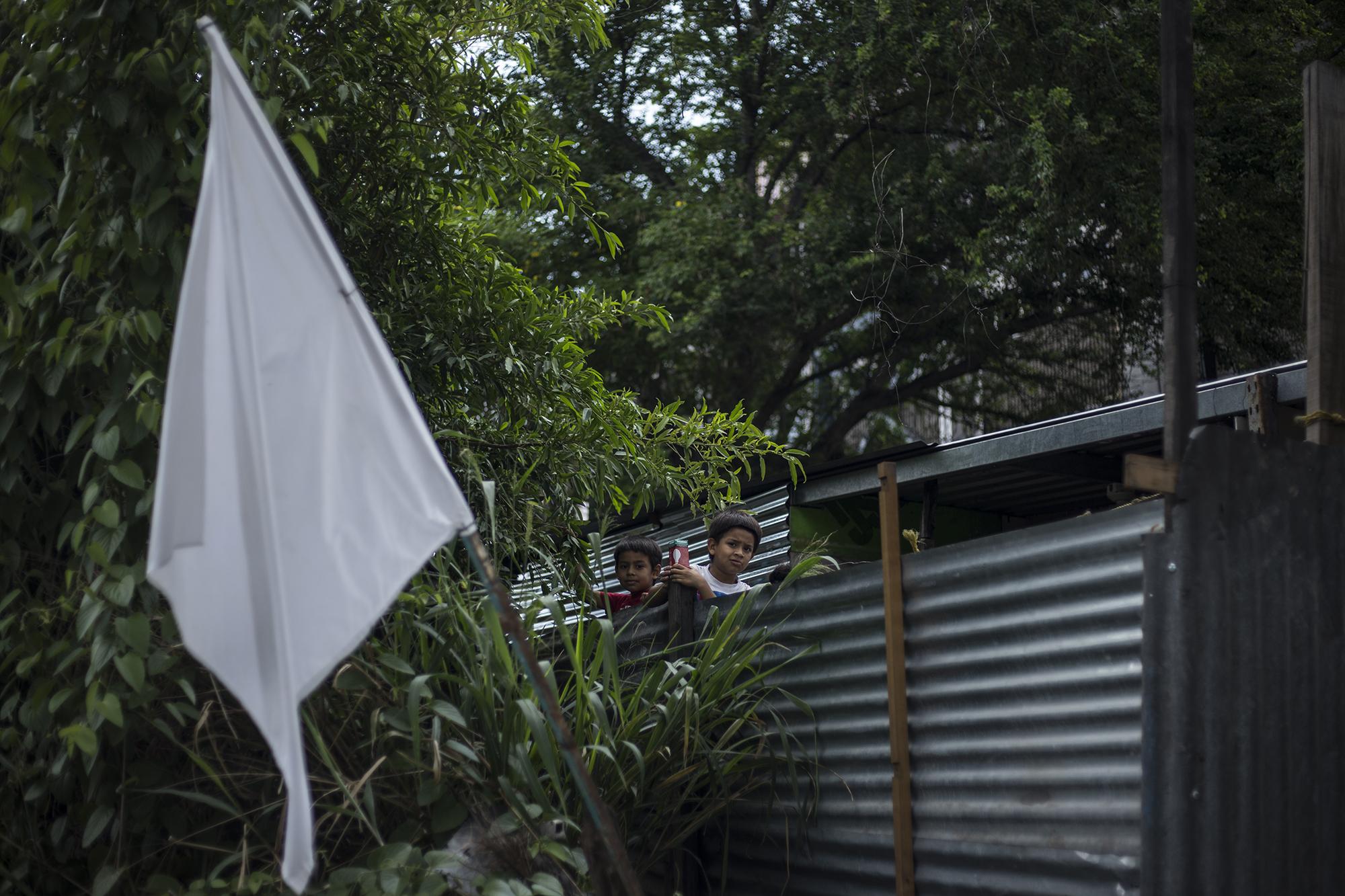 Los hermanos, Kevin y Diego López, de seis y siete años respectivamente, cuentan, desde las láminas que sirven de muro perimetral de su casa, los vehículos que circulan sobre el bulevar del Ejército. Es la entrada a la comunidad 15 de Marzo, establecida desde el año 2008, entre los límites de los municipios de San Salvador y Soyapango. Un conjunto de 125 casas de lámina vieja y madera en un predio baldío. El hambre arrecia en esta comunidad, habitada por al menos 500 personas; la mayoría, vendedores ambulantes que no han vendido por más de 70 días. Foto de El Faro: Víctor Peña.