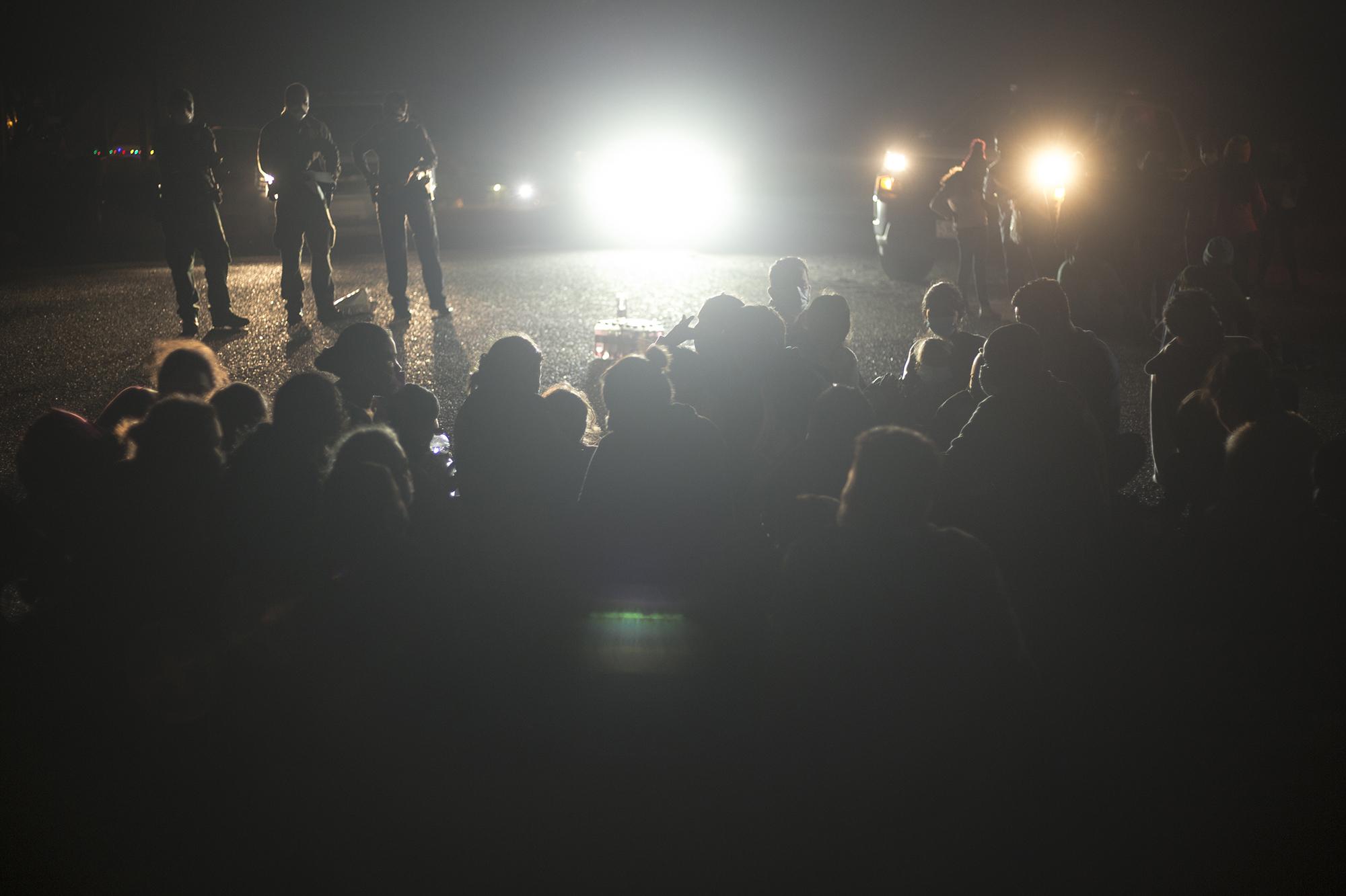 Dozens of minors take shelter in their parents’ arms as a large group sits in the dirt. They wait for hours to formally register with U.S. officials in Roma, Texas. Neighbors approach along the river to leave water and food.