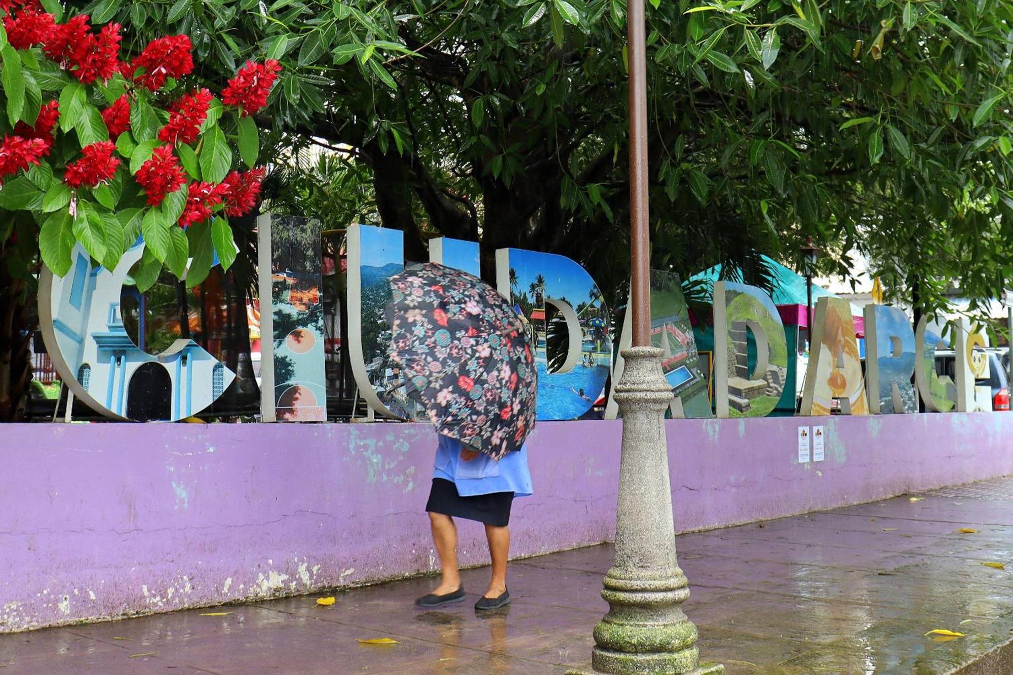 Una señora se protege de la lluvia –y del fotógrafo– mientras camina frente a las letras ya desteñidas que integran el nombre de la ciudad, en el parque Central de Ciudad Arce. Foto de El Faro: Roberto Valencia.