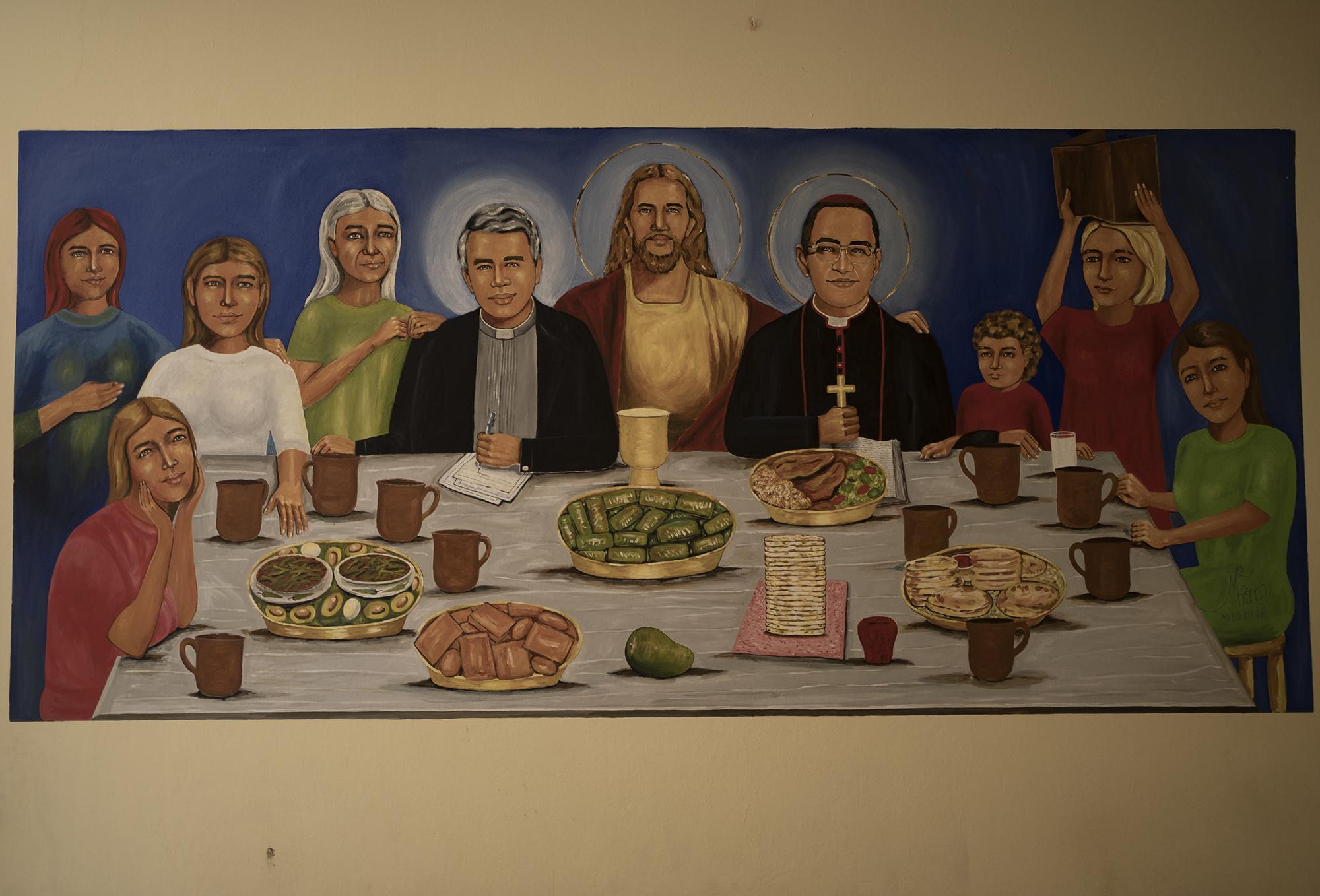 Representación de la Santa Cena en una pared de la iglesia católica de El Paisnal, donde se incluye a Rutilio Grande y San Romero. En la mesa, pupusas, tamales, marquesote, frijoles con aguacate y un mango.