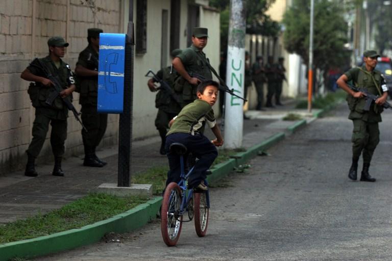 Un niño pasa con su bicicleta cerca de agentes de la Policia Nacional Civil (PNC) y soldados del Ejercito de Guatemala en Ciudad de Sol, en la periferia sur de Ciudad de Guatemala. Cinco soldados están procesados por propinar una golpiza y someter a abuso de autoridad a un adolescente y a un adulto que, según las propias fuentes militares, están relacionados a pandillas. Foto AFP