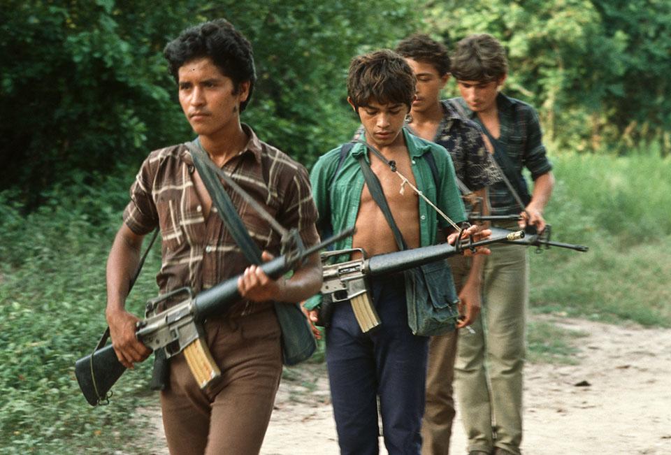 Imagen tomada en 1986 en un campamento guerrillero ubicado en el departamento de Usulután. Cuatro jóvenes, casi niños, patrullan con sus M-16/A1, el fusil que más proliferó entre los combatientes aglutinados bajo la sigla FMLN. Foto Giovanni Palazzo / MUPI.